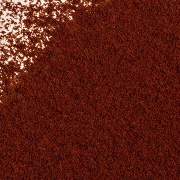 A close-up of Whole Spice Inc.'s Cloves Ground Organic—a fine, reddish-brown spice—spread across a white surface with some loose granules scattered at the top left.
