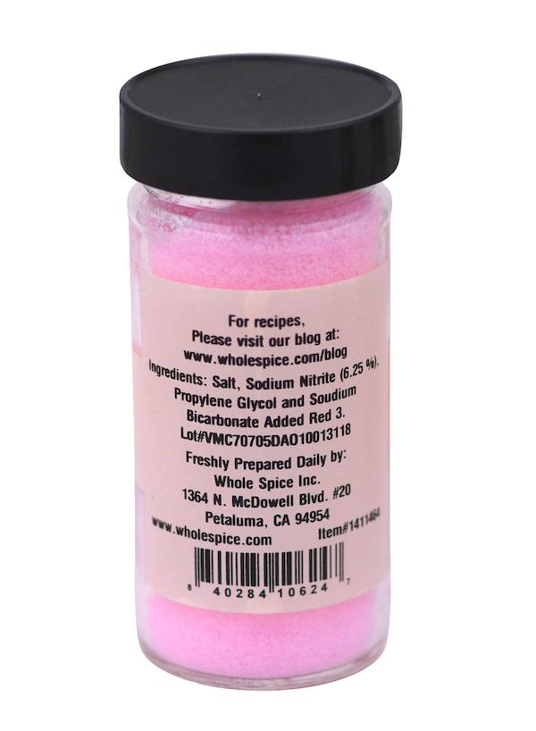 A clear jar of Whole Spice Inc. Curing Salt filled with pink curing salt for pickling meats. It has a black screw-on lid and a beige label displaying ingredients, preparation info, the website, and a barcode in black text.