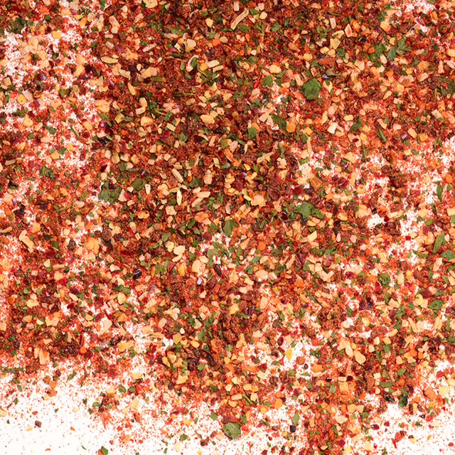 A close-up of Whole Spice Inc.’s Chipotle Lime Black Beans Seasoning—a vibrant blend of red pepper flakes, green herbs, and orange spices—scattered on white, ideal for adding bold flavor to black bean salsa.