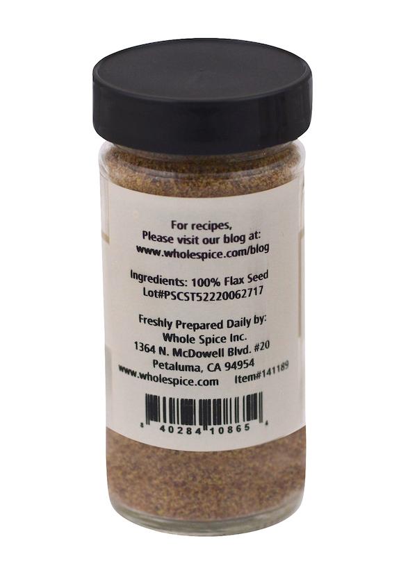 A clear jar with a black lid contains ground flax seeds by Whole Spice Inc. The label states "Flax Seeds Ground," ingredient: 100% flax seed, and includes company info, location, website, and a barcode at the bottom.