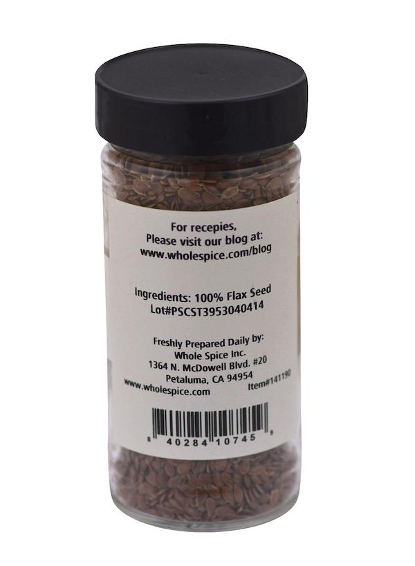 A clear plastic jar of Whole Spice Inc. Flax Seeds with a black lid and a back label listing 100% heart-healthy flax seeds as the ingredient, plus company and blog info. The jar contains brown flax seeds.