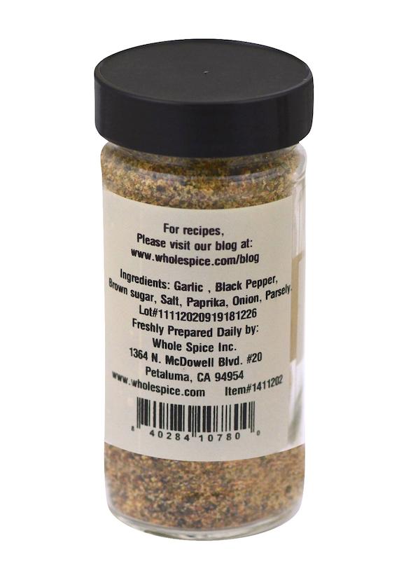 A clear spice bottle with a black lid features a label for Whole Spice Inc.'s Garlic Pepper Seasoning—listing garlic, black pepper, brown sugar, salt, paprika, onion, and parsley—ideal for meat. Company and website info appear on the label.