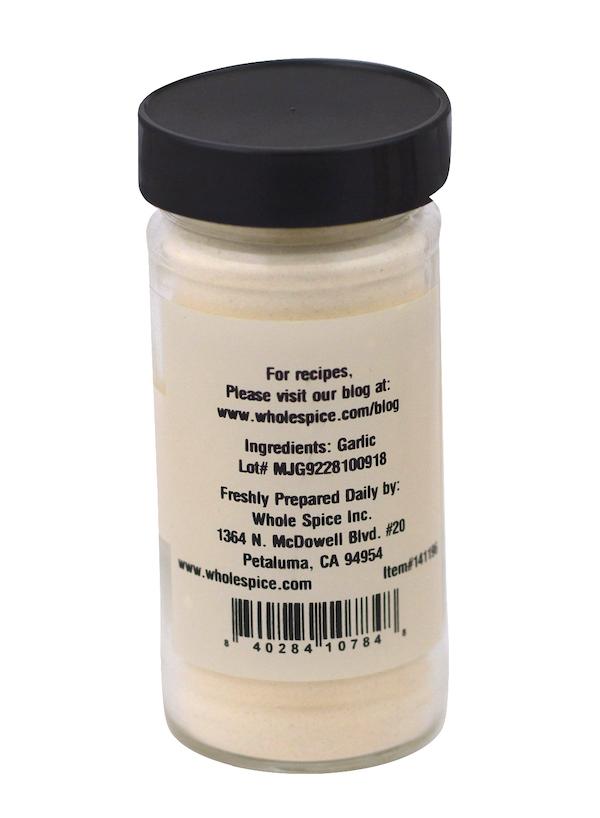 A clear spice jar with a black lid contains Whole Spice Inc. Garlic Powder, made from dehydrated garlic. The label lists garlic as the ingredient, along with the company’s address, website, and a barcode.