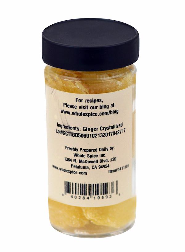 A clear spice jar from Whole Spice Inc. with a black lid contains Ginger Crystallized. The upright jar's label lists ingredients, company info, a recipe website, and has the barcode at the bottom.