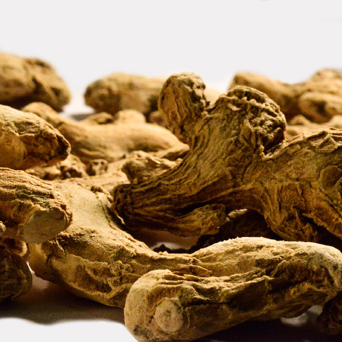 A close-up of Ginger Whole by Whole Spice Inc. shows several dried roots with rough, wrinkled textures and earthy brown color, scattered on a white surface with clearly visible twisted shapes.