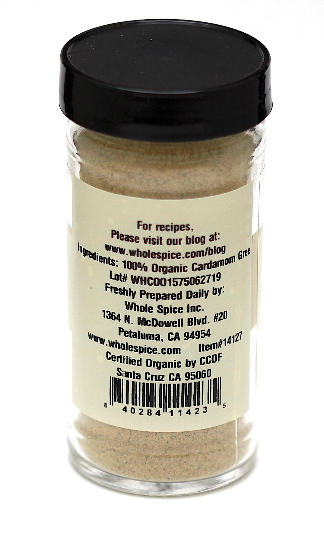 A clear spice jar with a black lid from Whole Spice Inc. contains pale brown Green Cardamom Powder Organic. The label lists 100% ground cardamom as the ingredient, manufacturer info, certification details, and a barcode at the bottom.