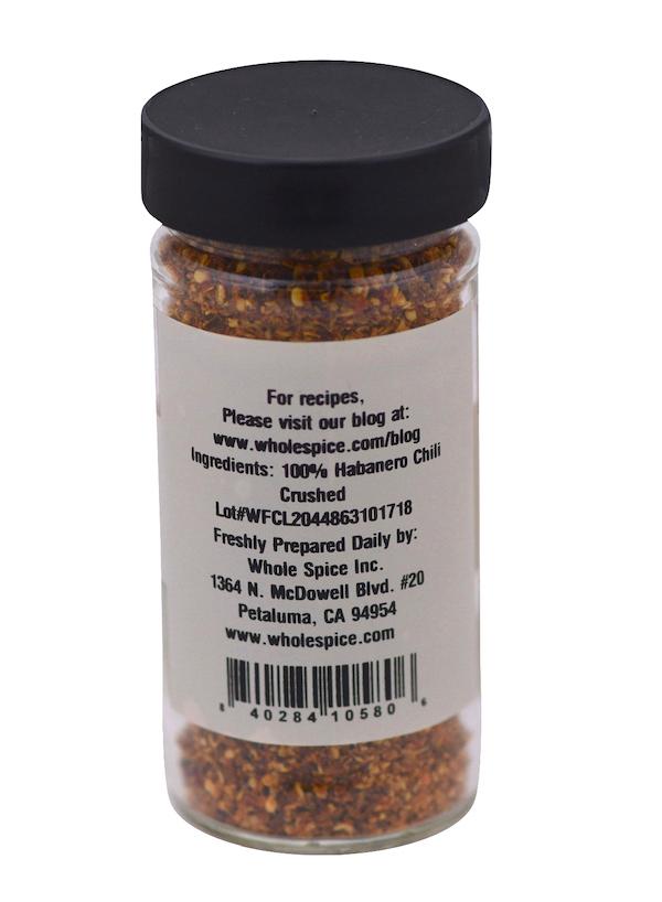 A clear spice jar with a black lid contains vibrant crushed red and orange hot chili pepper. The label reads "Habanero Chili Crushed" by Whole Spice Inc., listing ingredients, company info, and Scoville heat units for intense heat.