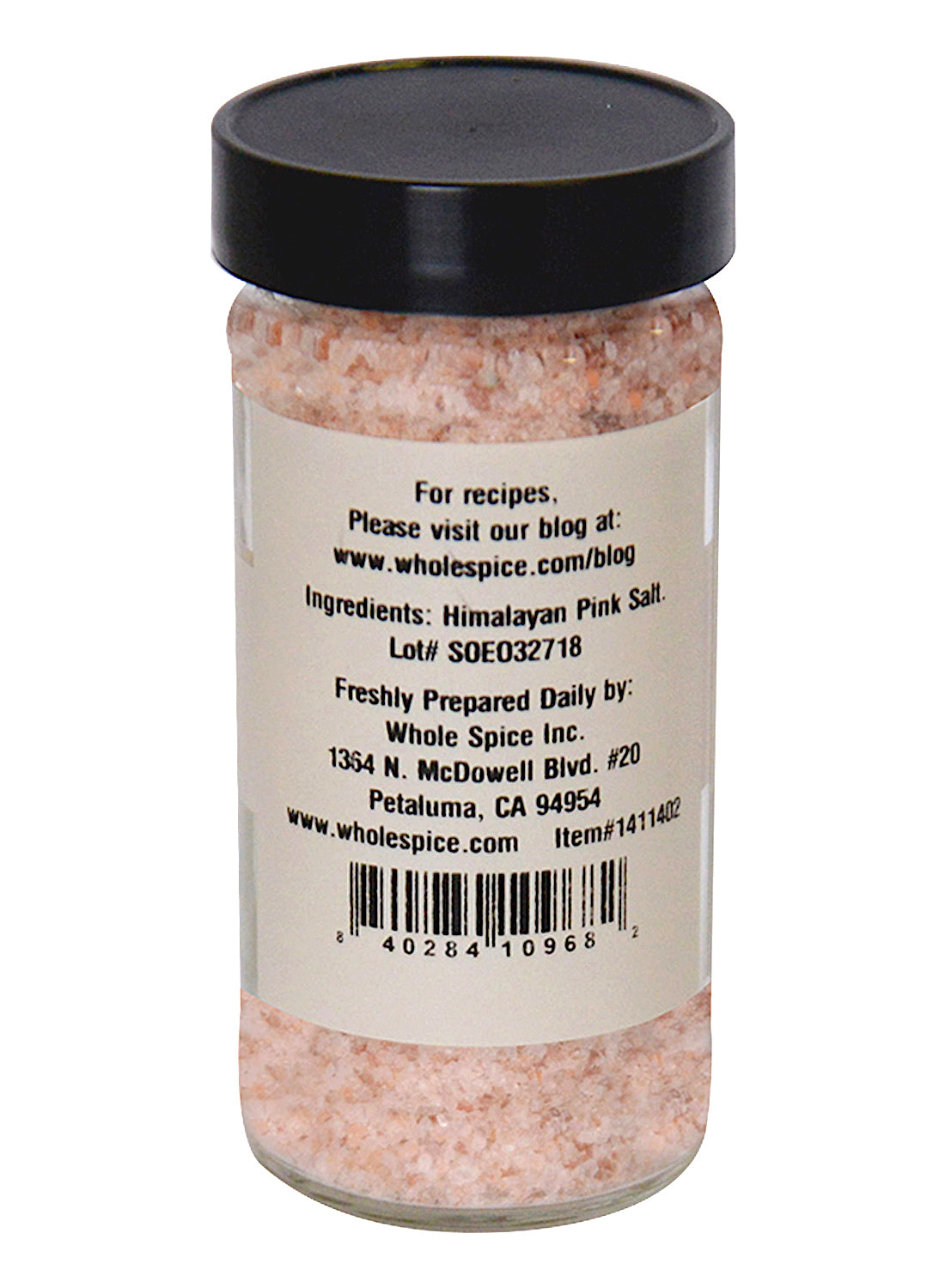 A clear glass jar with a black lid contains Himalayan Pink Salt Medium #2 (2-3mm) by Whole Spice Inc. The label lists ingredients, company info, barcode, website, lot number, and contact address. Prized for its trace minerals.
