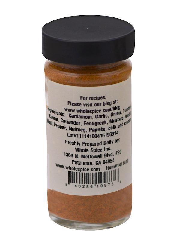 A clear spice jar with a black lid contains Whole Spice Inc. Hot Curry Powder, an orange-red Indian blend featuring turmeric and paprika. The label shows ingredients, recipes website, company address, and barcode.