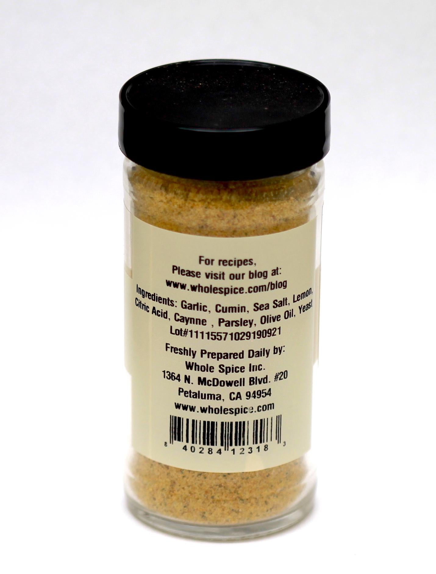A clear spice jar from Whole Spice Inc. labeled "Hummus Spice Blend" features a black lid and contains yellowish powder with ingredients like garlic, cumin, sea salt, and lemon—ideal for adding flavor to hummus. Manufacturer details are provided.