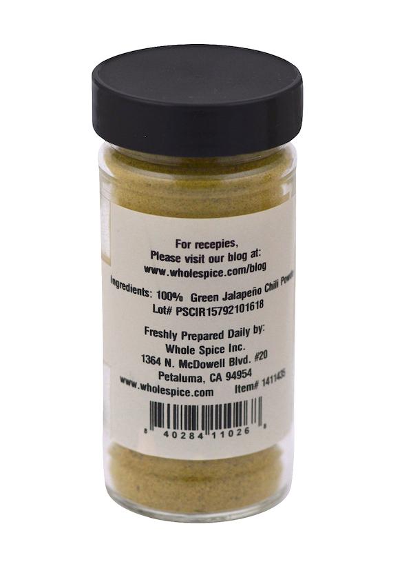A clear spice jar with a black lid contains Jalapeño Green Chili Powder by Whole Spice Inc. The label shows the product name, brand, supplier’s address, website, and a barcode at the bottom. The powder inside is vibrant green.
