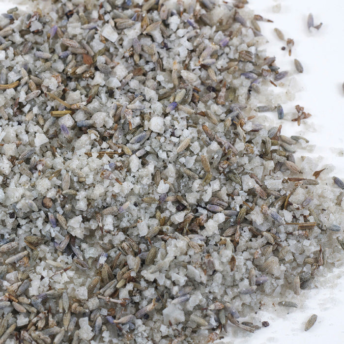 A close-up of Whole Spice Inc. Lavender Salt, featuring a blend of coarse sea salt and dried lavender buds scattered on a white surface.