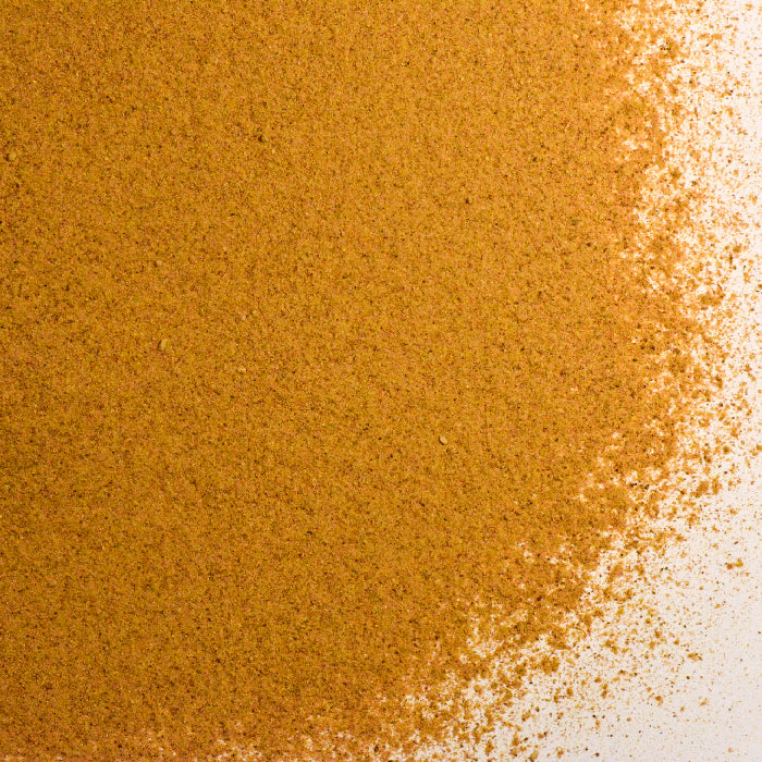 A close-up of Whole Spice Inc. Maca Powder, a fine brown superfood, spread in a pile with uneven edges on a white surface.