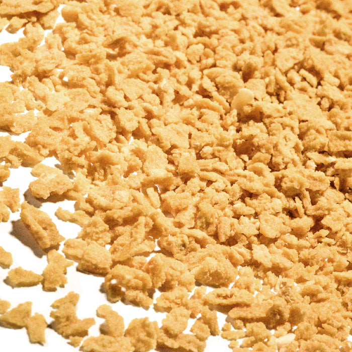 A close-up of Maple Flakes by Whole Spice Inc.—crispy golden-brown cornflakes mixed with crunchy maple flakes—scattered on a white surface.