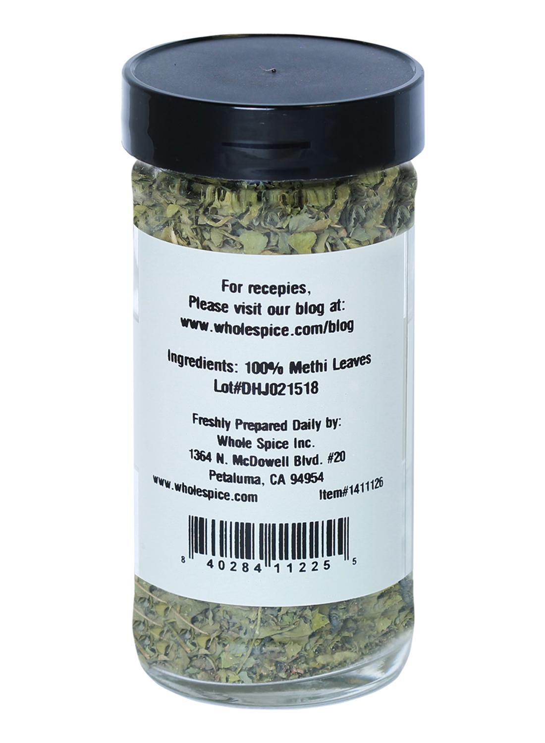 A clear spice jar of Whole Spice Inc. Methi Leaves is filled with dried methi, ideal for Indian dishes. The white label lists "Ingredients: 100% Methi Leaves" with a website, lot number, address, barcode, and a closed black lid.
