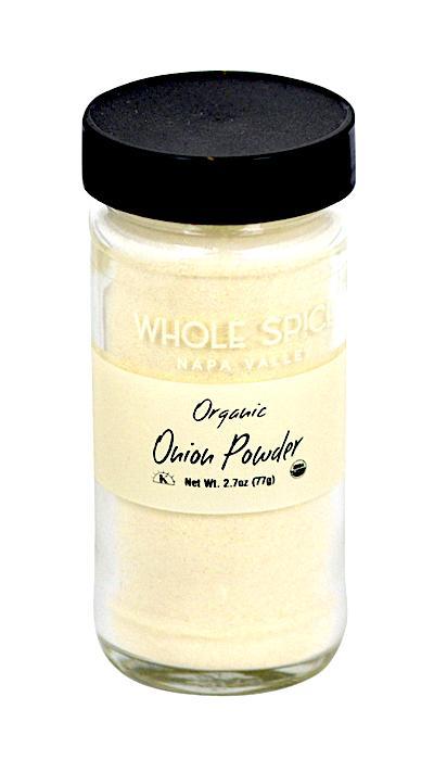 A clear glass jar with a black lid holds light onion powder labeled Whole Spice Inc. Onion Powder Organic, made from organic onion and blended with a natural anti-caking agent. Net Wt. 2.7 oz (77g).
