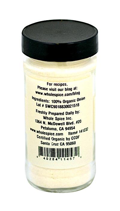 A clear glass jar with a black lid holds Whole Spice Inc. Onion Powder Organic, made with a natural anti-caking agent. The white label in black text lists ingredients, company info, barcode, and website for recipes.