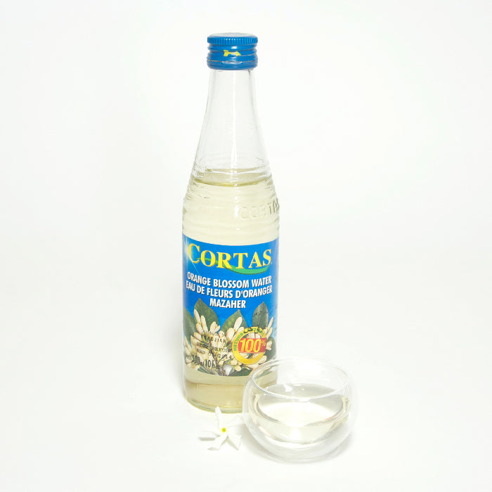 A clear glass bottle of Whole Spice Inc. Orange Blossom Water 10 oz stands beside a small bowl of citrusy liquid and white orange blossoms, ideal for enhancing Middle Eastern pastries, all set against a white background.