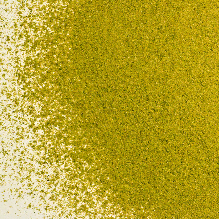 A close-up of Whole Spice Inc. Oregano Powder, greenish-yellow in color, scattered densely on the right and fading lightly to the left across a white background.