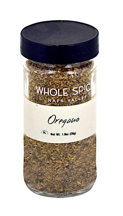 A clear glass jar with a black lid holds dried Greek oregano. The label reads Whole Spice Inc. Oregano(Greek), Net Wt. 1 oz (28g).