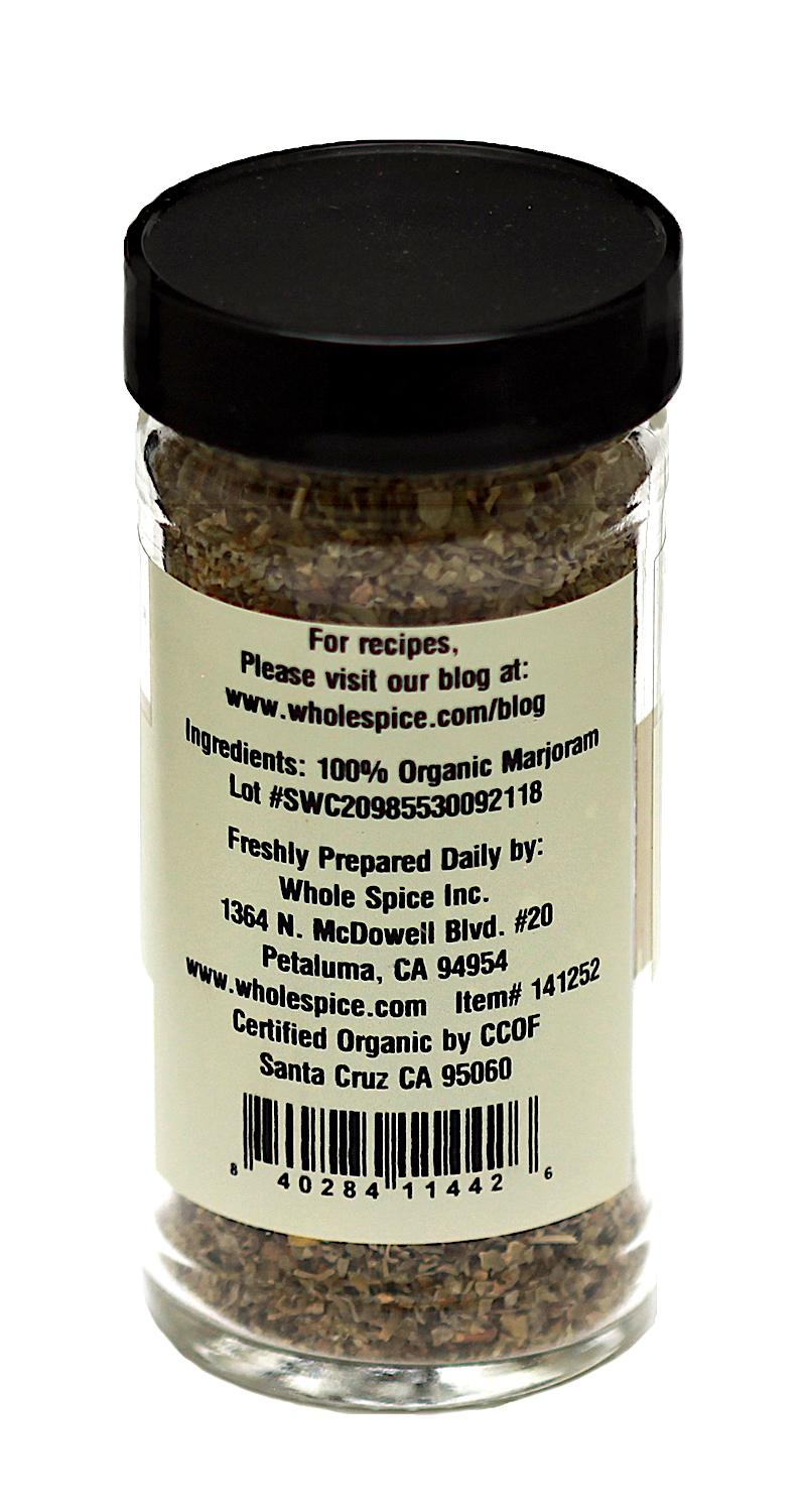 A clear spice jar with a black lid contains Marjoram Leaves Organic by Whole Spice Inc., labeled 100% organic with producer details, website, address, and certification for premium organic herbs.