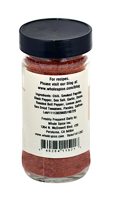 A clear spice jar with a black lid features a white label for Paella Spice Mix by Whole Spice Inc., listing Mediterranean spices such as chili, smoked paprika, and black pepper, plus preparation details and contact information.