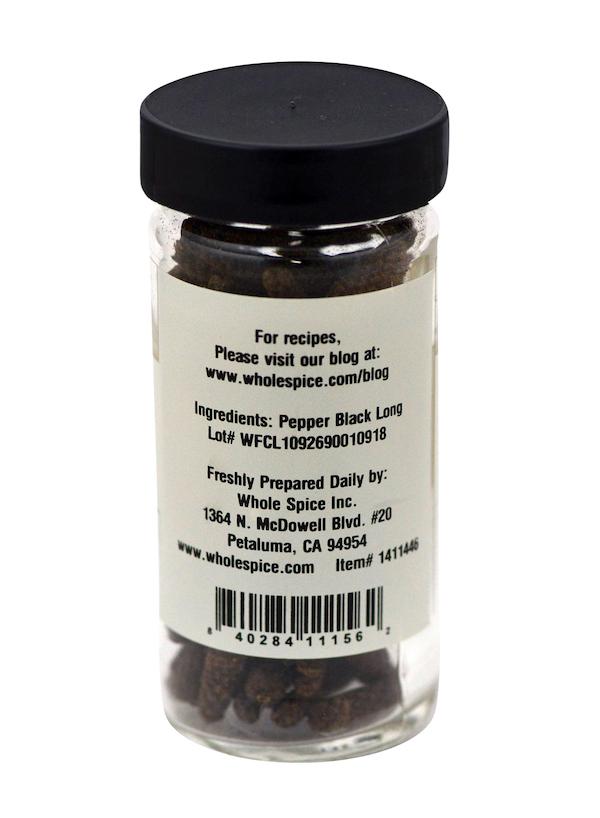 A clear spice jar with a black lid holds Pepper Black Long from Whole Spice Inc. The label includes ingredients, company info, barcode, lot number, and a website for recipes.
