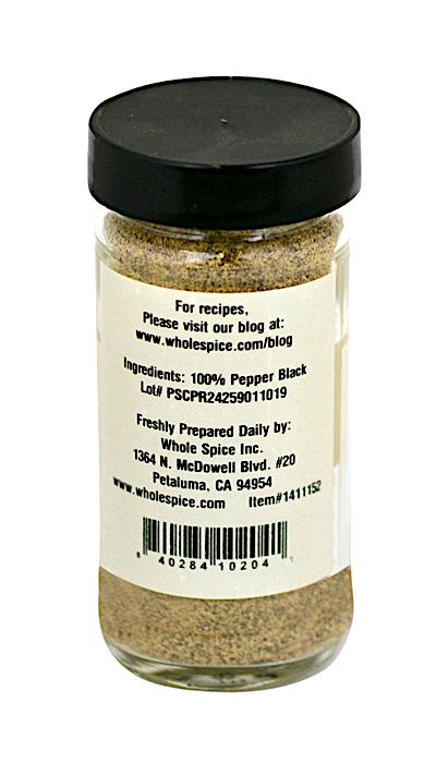 A clear glass jar with a black lid contains Pepper Black Powder by Whole Spice Inc. The label lists 100% black peppercorns as the ingredient, along with company info, address, lot number, and a barcode.