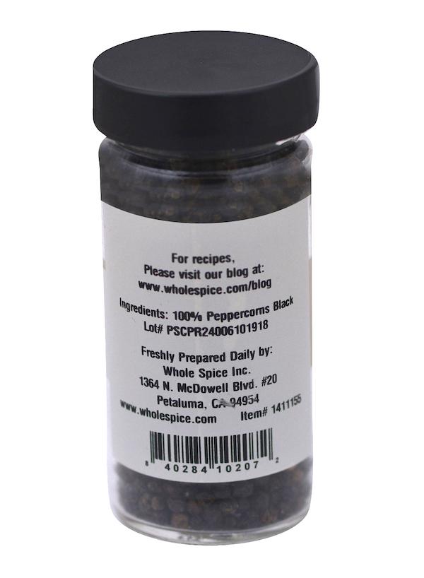 A clear plastic jar with a black lid contains Peppercorns Black from Whole Spice Inc.—100% black peppercorns, with contact info and website listed on the label.