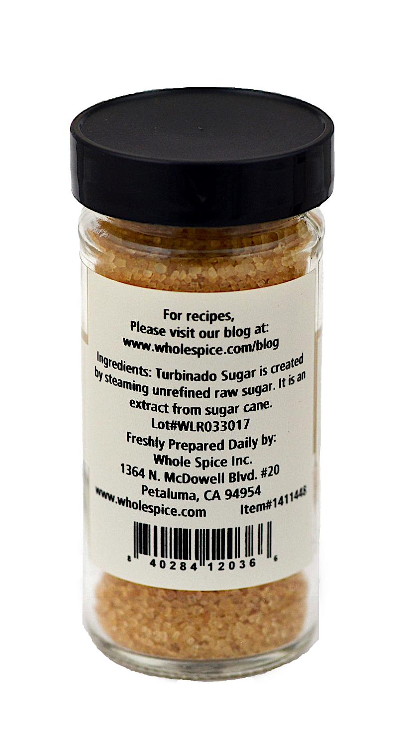 A glass jar with a black lid contains Turbinado Sugar from Whole Spice Inc. The label provides ingredients, contact info, and manufacturer details. Tan sugar crystals are visible through the jar.