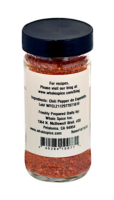 A clear glass jar with a black lid from Whole Spice Inc. contains crushed Piment d'Espelette; the label lists the ingredient, manufacturer info, and a blog link for recipes.