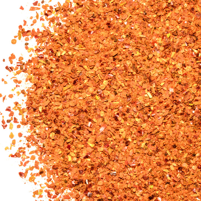 Close-up of Whole Spice Inc.'s Ras el Hanout Coarse Ground, a vibrant Moroccan spice blend, with scattered orange, red, and yellow flakes on a white background.