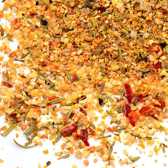 A close-up of Whole Spice Inc. Rosemary Green Olives Seasoning scattered on a white surface, showing visible bits of lemon peel, dried herbs, chili flakes, and spices in vibrant yellow, orange, green, and red hues.