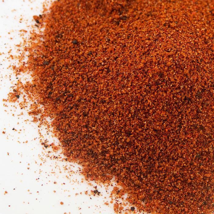 A close-up of Whole Spice Inc. Rib Rub, a reddish-brown ground spice blend scattered on a white surface, perfect for adding bold barbecue flavor to grilling or as a savory rib rub.