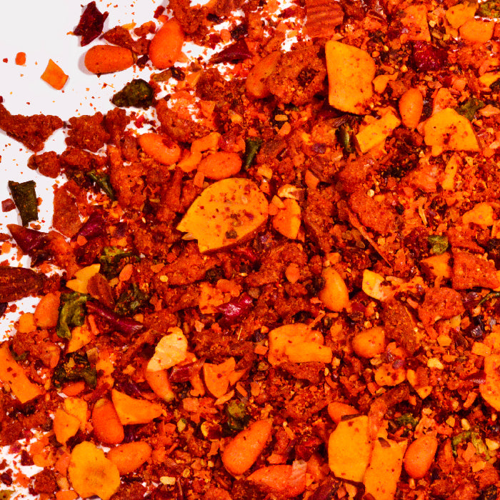 A close-up of Whole Spice Inc.'s Moroccan Harissa Rice Seasoning, showcasing vibrant orange and red hues with visible dried herbs, seeds, and crushed nuts scattered on a white surface.