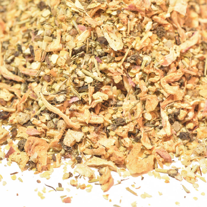 A close-up of Whole Spice Inc. Roast Beef Rub shows a blend of dried roots, herbs, and bark pieces in light brown and beige tones, evoking the aromatic seasoning mix scattered on a white surface.