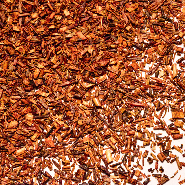 A close-up of Whole Spice Inc. Rooibos Tea leaves, caffeine-free and rich in antioxidants, scattered on a white surface to display their reddish-brown hue and finely chopped texture.
