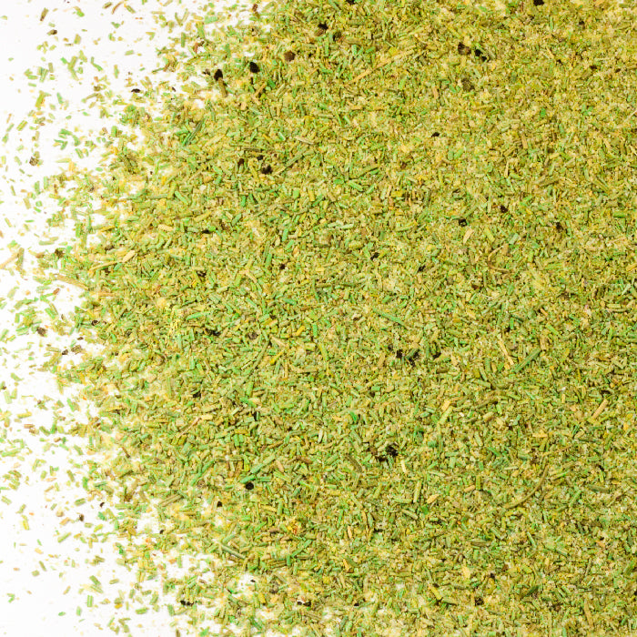 Close-up of Whole Spice Inc. Rosemary & Garlic Rub, a finely chopped blend of dried green herbs spread on a white surface—perfect for enhancing the flavor of meat dishes.
