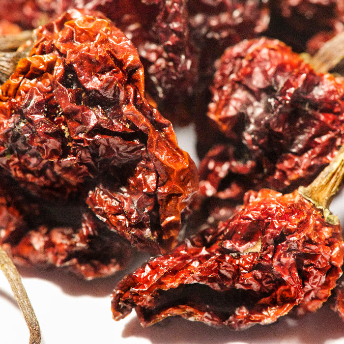 Close-up of Trinidad Moruga Scorpion Pepper Whole by Whole Spice Inc., showcasing several wrinkled red chilies with brown stems, arranged on a white surface.