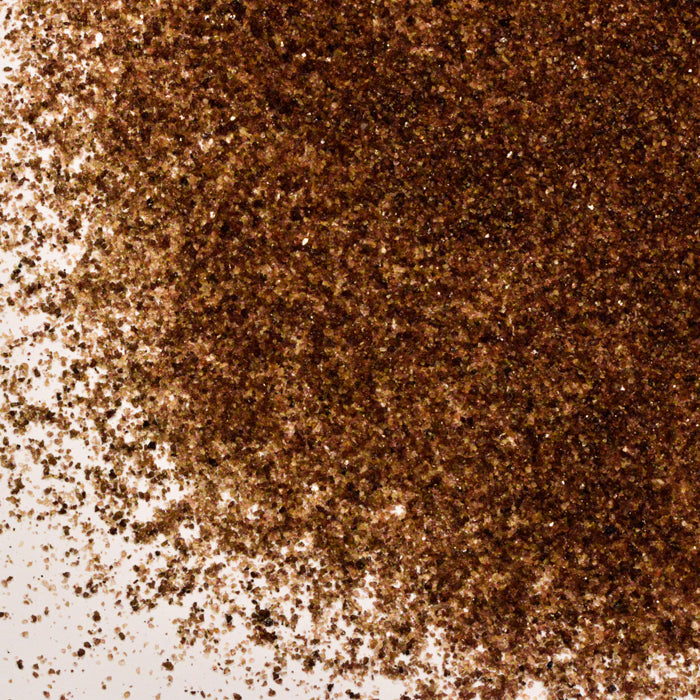 A close-up shows Alderwood Smoked Sea Salt by Whole Spice Inc., its coarse grains piled densely in the upper right and scattered more sparsely toward the lower left on a white surface.