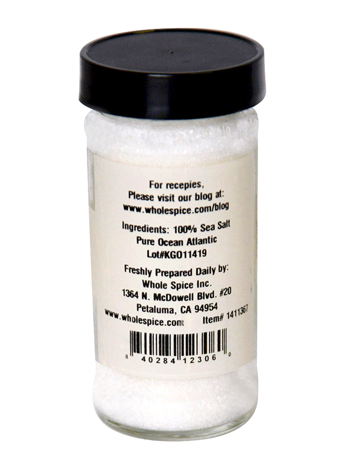 A clear glass jar with a black lid contains Sea Salt Fine by Whole Spice Inc. The label displays ingredients, contact details, and a barcode.