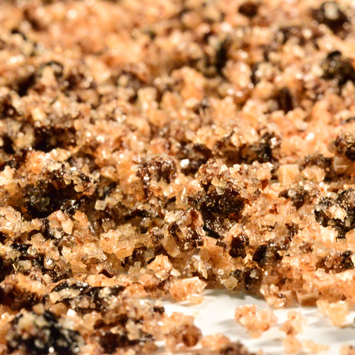 A close-up view of Olive Salt by Whole Spice Inc. shows coarse brown crystals mixed with dark clumps and a sprinkle of black olive sea salt, all scattered on a light surface.
