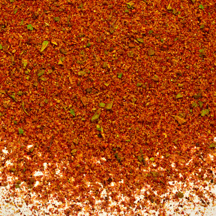 A close-up of finely ground red chili powder and dried herbs spread on a white surface—Whole Spice Inc. Taco Seasoning, an authentic Mexican blend perfect for tacos.