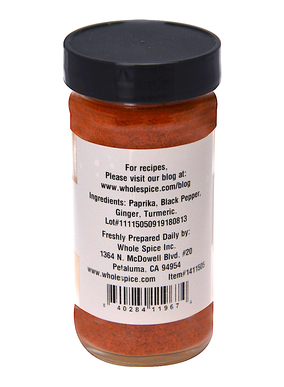 A clear jar of Tagine Spice by Whole Spice Inc. with a black lid contains red-orange powder. The label highlights North African spices—paprika, black pepper, ginger, turmeric—and features company info and a barcode at the bottom.