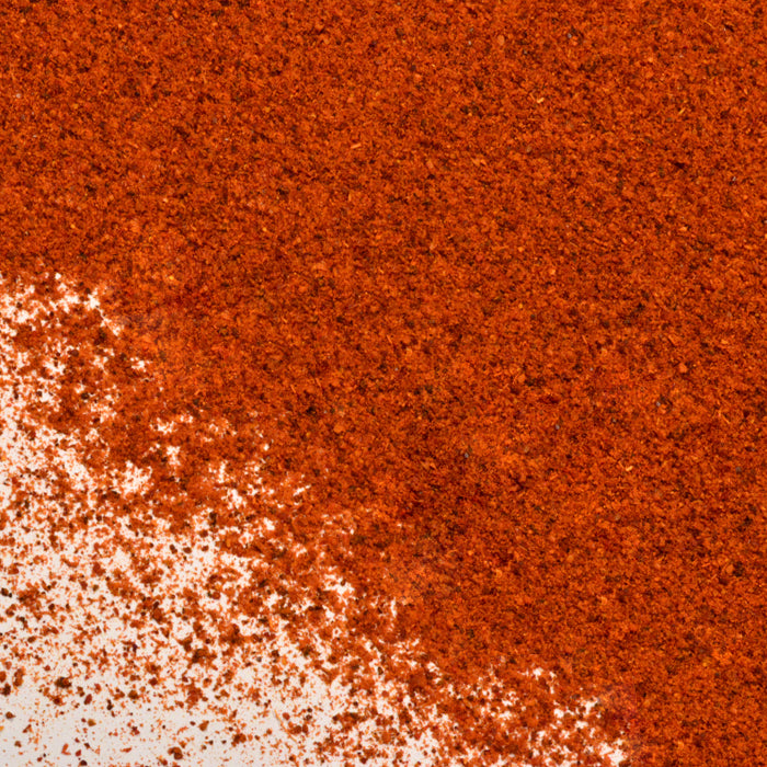 A close-up of finely ground, bright red-orange Tagine Spice by Whole Spice Inc., spread unevenly on a white surface.