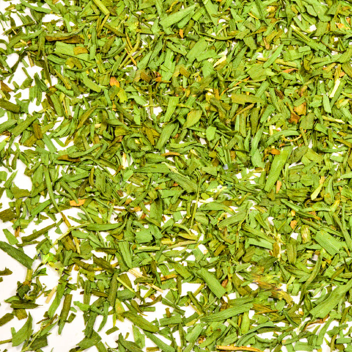 Close-up of Whole Spice Inc. Tarragon—dried, chopped green herbs with thin, irregularly shaped leaves scattered on a white surface, classic in French cuisine.