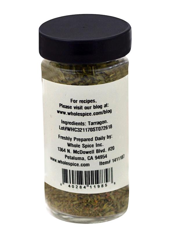 A glass spice jar from Whole Spice Inc., labeled Tarragon, features a black lid and contains dried tarragon, a classic French herb. The label includes ingredient info, manufacturer details, address, and website for recipes.
