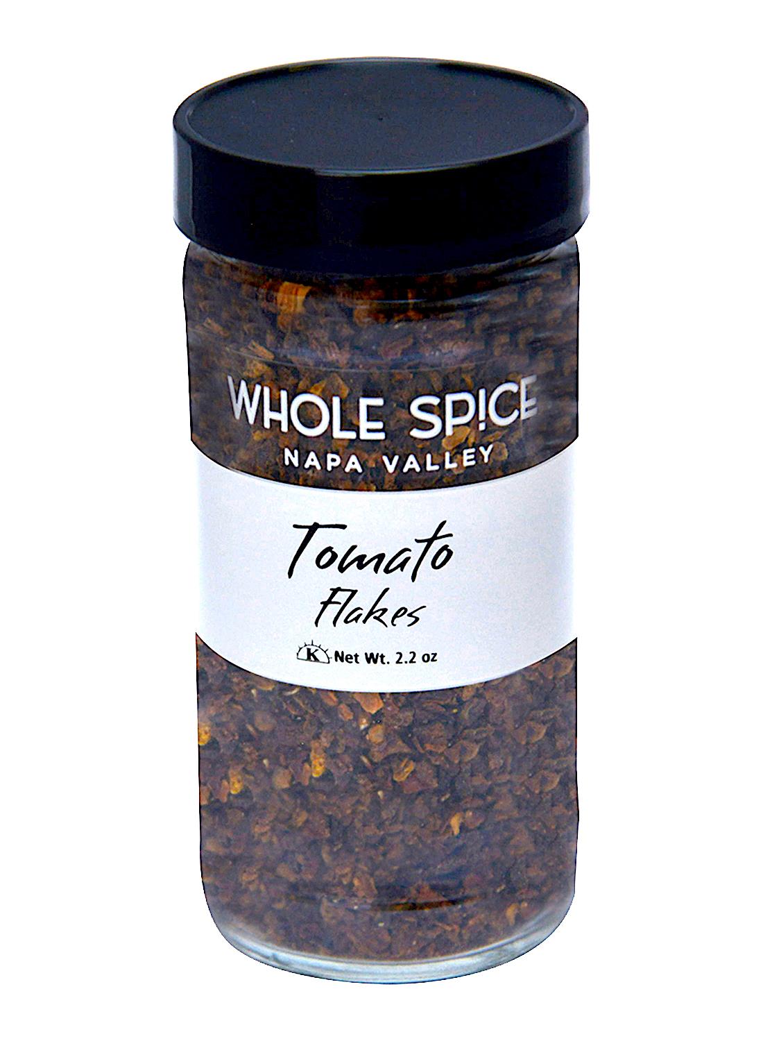 A clear glass jar with a black lid labeled Whole Spice Inc. Tomato Flakes contains dried tomato flakes free from sulfur dioxide, net weight 2.2 oz.