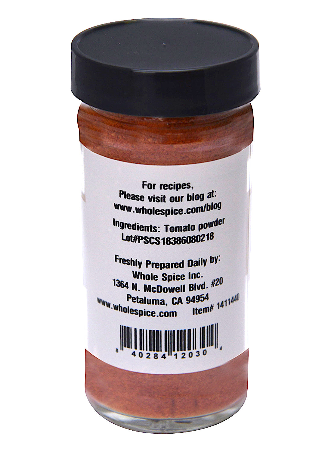 A clear spice jar with a black lid from Whole Spice Inc. contains Tomato Powder made from dehydrated tomatoes. The label lists ingredients, lot number, company contact info, recipe website, and has a barcode at the bottom.