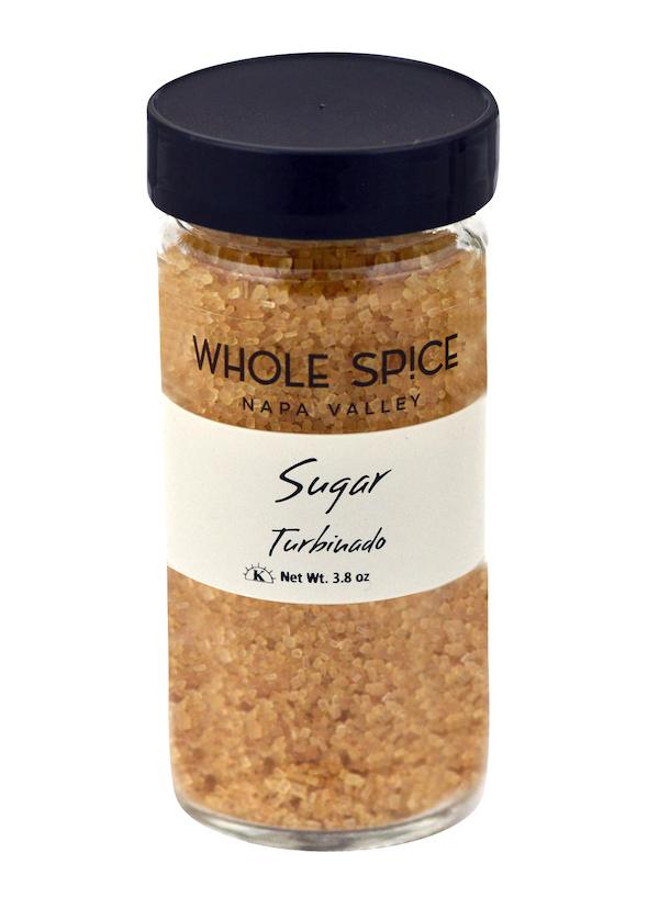 A clear glass jar with a black lid labeled Whole Spice Inc. Turbinado Sugar, containing coarse, light brown unrefined sugar crystals. Net weight: 3.8 oz.